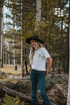 Girl standing in a forest wearing a unisex graphic-tee that is white with a small yellow smiley face wearing a cowboy hat with the word howdy below it on the left side of the tshirt. She is also wearing a black wide brim hat and dark blue jeans.