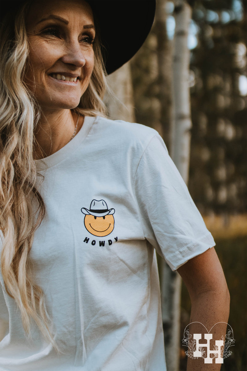 Girl wearing a white unisex tshirt with a yellow smiley face wearing a cowboy hat with the word howdy below it. The smiley face is small and on the left side of the tshirt. 