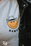 Close up of graphic on white shirt. Graphic is small yellow smiley face wearing a cowboy hat with the word howdy below it.