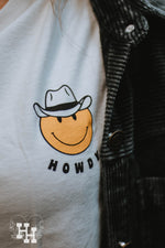 Close up of graphic on white shirt. Graphic is small yellow smiley face wearing a cowboy hat with the word howdy below it.