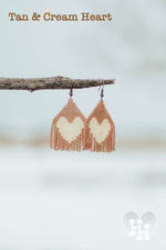Set of dangly seed bead earings hanging on a stick on a snowy day. Earings are tan with a cream heart.