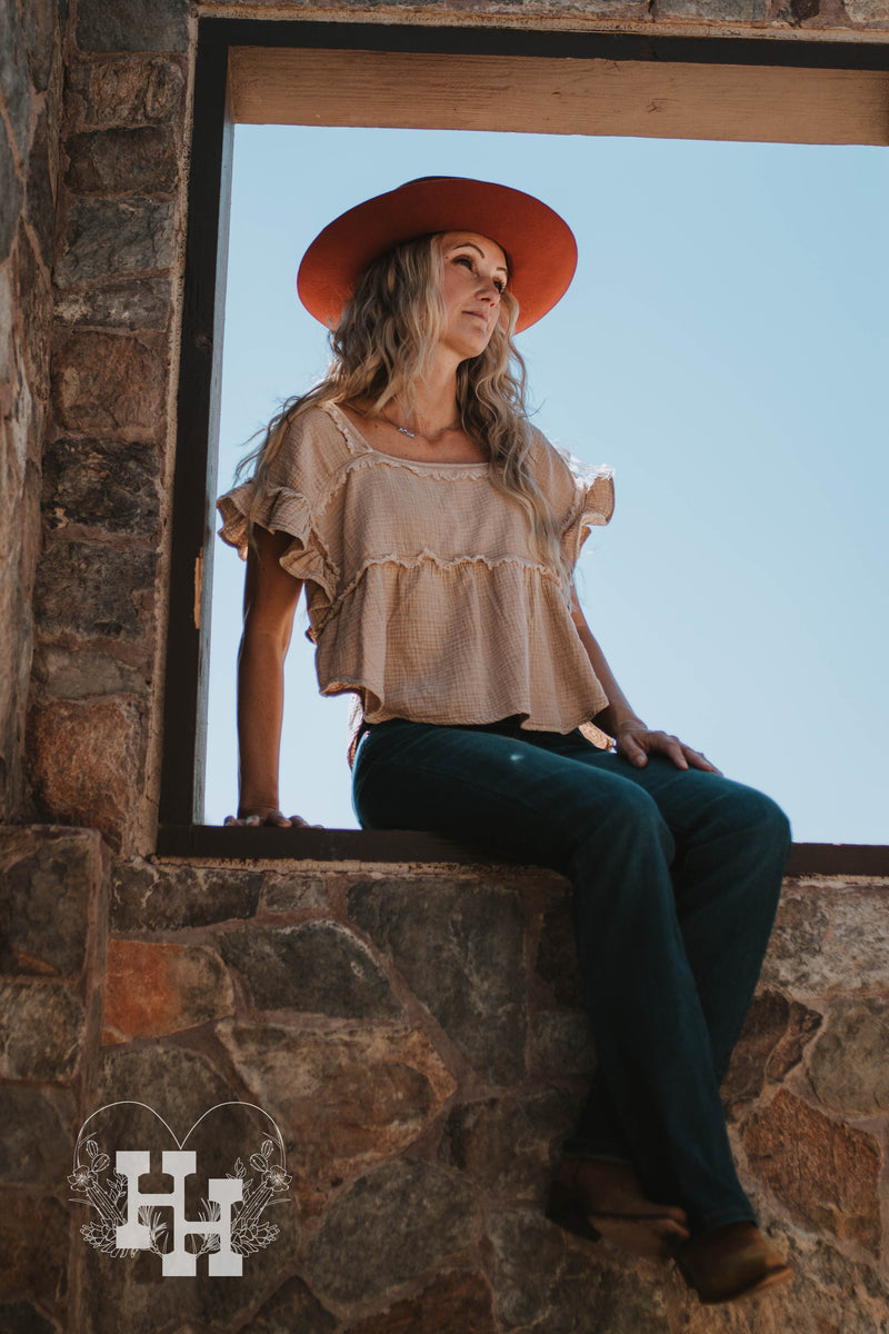 Girl sitting in a window seal of an old rock house. She is wearing a tan loose fitting crop top with a crinkle texture and unfinshed seems and ruffled sleeves. She is wearing it with dark blue jeans a burnt orange wide brim hat and boots.