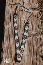 Handmade seed bead hat band laying accross a weathered wooden beam. Band is White, Turquoise, brown, gold and black in an X and diamond pattern. The end have black leather straps to add it on to a wide brim or western style hat.