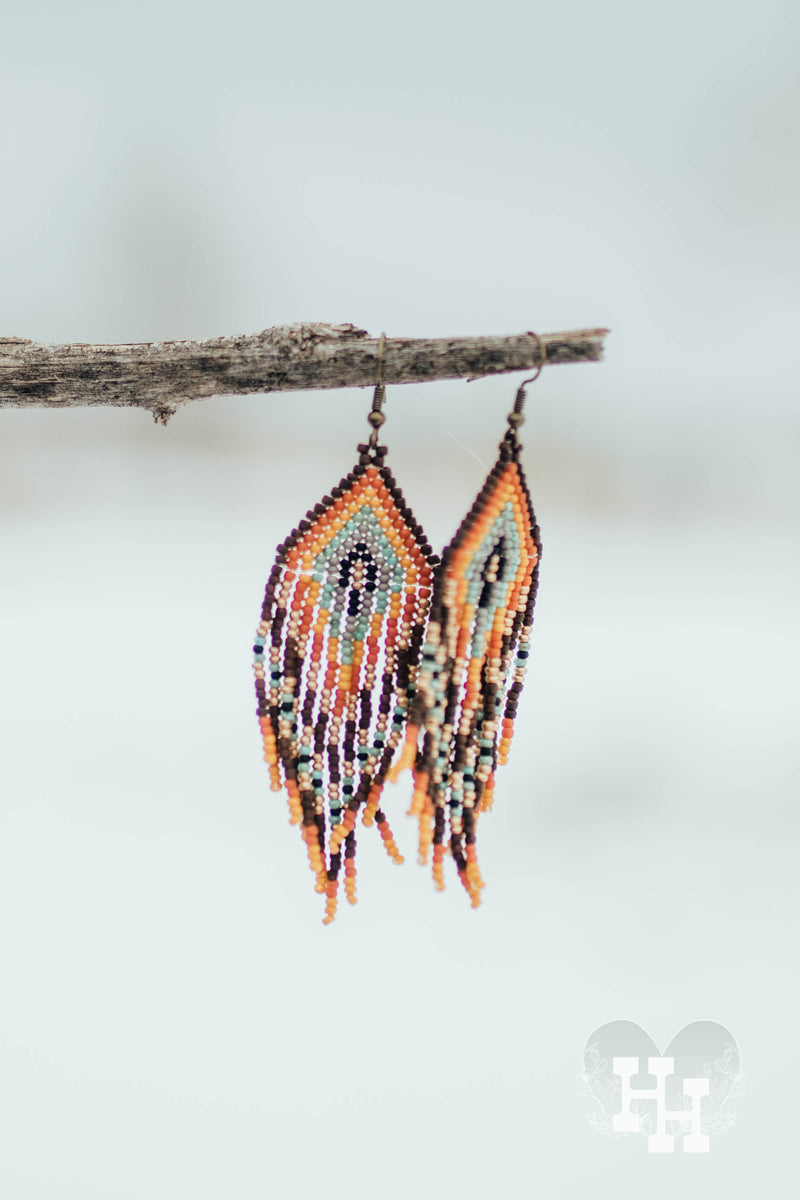 Set of seed bead earrings hanging on a stick on a snowy day. Earrings are traingle rainbow pattern with same pattern dangling below. Brown, Orange, Yellow, Turquoise, Gray, Black and Gold.