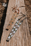 Two handmade seedbead hat bands laid accross a weathered wooden beam. Top band is yellow, white, tan, and green with a x and diamond pattern. It also had a brown leather strap on the ends put it around a hat. Bottom hat band is white, turquoise, black and brown, with same x and diamond pattern. It has black leather straps on the ends for putting it around a wide brim or western hat.