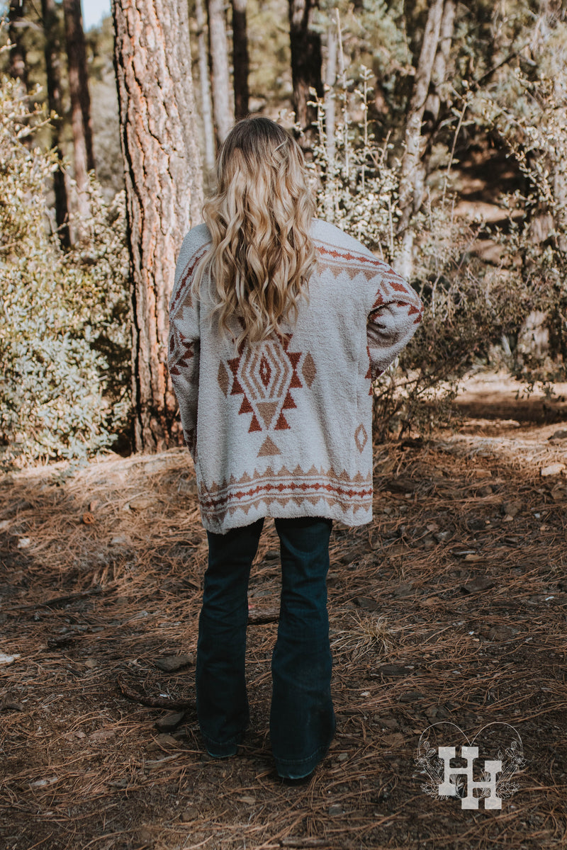 Back view of girl wearing aztec printed cardigan sweater. Cardigan sweater is long enough to cover her butt. Most of the sweater is offwhite with the aztec pattern being burnt orange and tan.
