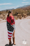 Girl standing in middle of dirt road wearing cowboy boots, denim shorts, and a red tshirt with a Bald Eagle head wearing sunglasses and the word Merica accross the top in black. The girl also has the american flag wrapped around her.