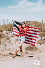 Girl holding American flag up behind her. She is wearing a red tshirt with black logo on it, log is a bald eagle head wearing sunglasses and the word Merica above it. She is also wearing denim shorts and cowboy boots.