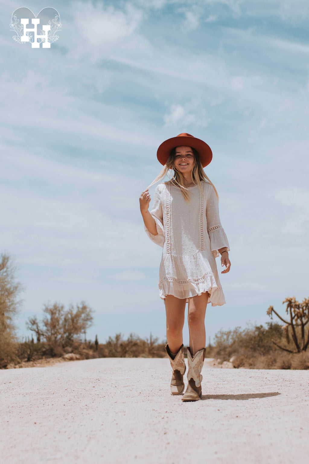 Girl walking down dirt road in a cream color mid thigh length dress. Dress has a drop waist with 3/4 length sleeves that have belled details at the end. Fabric is swiss dotted. She is wearing the dress with boots and a burnt organe wide brim hat.